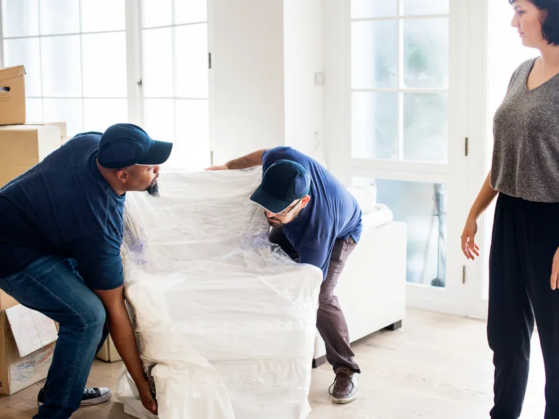 Uniformed movers wrapping a sofa while the client looks on